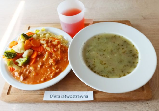 Taca z obiadem w diecie łatwostrawnej. Po lewej stronie znajduje się talerz z makaronem świderkami, gotowanymi warzywami (marchew, brokuły, kalafior) i sosem mięsno-pomidorowym. Po prawej – talerz z zieloną zupą jarzynową. W tle plastikowy kubek z czerwonym napojem owocowym