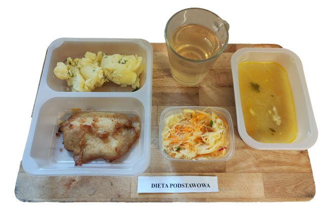 Taca z obiadem w diecie podstawowej: ziemniaki z koperkiem i panierowany filet z kurczaka w plastikowym naczyniu, obok surówka z kiszonej kapusty i marchewki w przezroczystym pudełku, zupa jarzynowa z kawałkiem ziemniaka i zieleniną oraz szklanka jasnego naparu (herbaty lub wody smakowej)