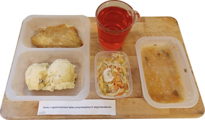 „Dieta z ograniczeniem łatwo przyswajalnych węglowodanów” Zestaw obiadowy na drewnianej tacy, składający się z porcji ziemniaków puree z koperkiem, panierowanego fileta rybnego, surówki z kapusty kiszonej z marchewką i jabłkiem, zupy jarzynowej oraz szklanki czerwonego napoju owocowego