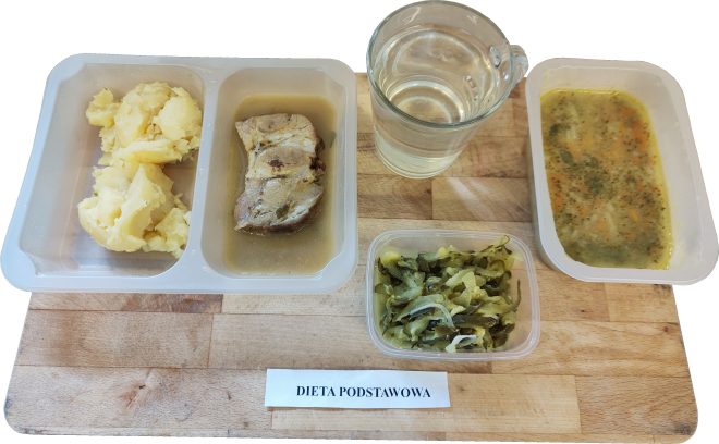 Na tacy znajduje się posiłek obiadowy oznaczony jako „Dieta podstawowa”: ziemniaki puree, duszony kawałek mięsa w sosie, surówka z ogórków kiszonych i cebuli w plastikowym pojemniczku oraz zupa jarzynowa. Do posiłku podano szklankę wody