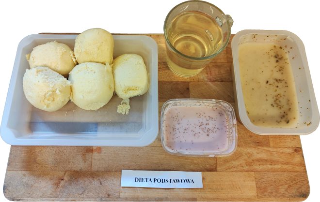 Zestaw obiadowy w diecie podstawowej podany na desce: porcja kilku ziemniaków w pojemniku, szklanka jasnego napoju, zupa krem w plastikowym pojemniku oraz deser mleczny w małym pojemniczku. Na desce widoczny napis Dieta podstawowa.