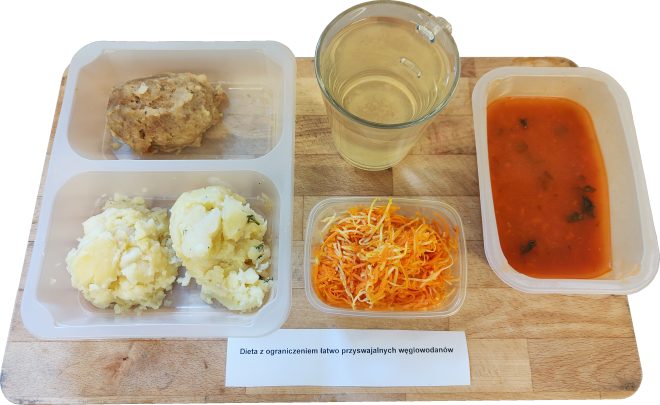 Na tacy drewnianej ułożony zestaw obiadowy. W plastikowym pojemniku podzielonym na dwie przegródki znajduje się kotlet mielony oraz porcja ziemniaków puree z koperkiem. Obok w osobnych pojemnikach: surówka z marchewki i selera, czerwony barszcz lub zupa pomidorowa z natką pietruszki oraz szklanka jasnego napoju. Na kartce pod tacą widnieje napis „Dieta z ograniczeniem łatwo przyswajalnych węglowodanów”