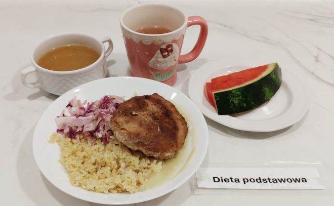 Na białym talerzu znajduje się porcja kaszy bulgur, kotlet schabowy w sosie oraz surówka z czerwonej kapusty. Do dania podano zupę w białej filiżance, herbatę w różowym kubku i kawałek arbuza na małym talerzyku. Na blacie widoczny jest napis „Dieta podstawowa”