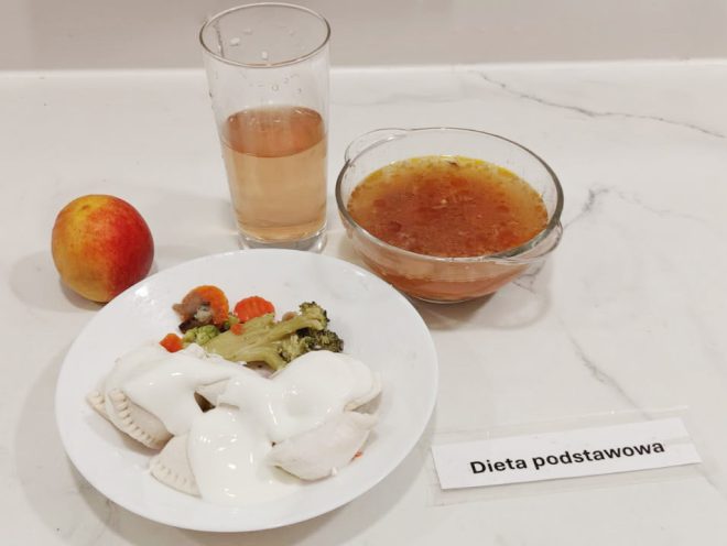Na białym blacie ustawiony jest zestaw obiadowy: talerz z pierogami polanymi białym sosem i porcją gotowanych warzyw (marchew, brokuł, kalafior), szklanka jasnego napoju, miseczka z zupą jarzynową o pomarańczowym kolorze oraz jabłko. Po prawej stronie karteczka z napisem „Dieta podstawowa”