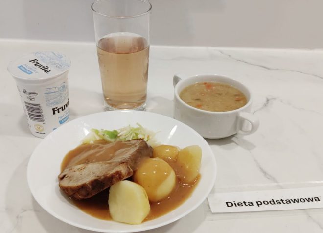 Na białym blacie stoi talerz z gotowanymi ziemniakami, kawałkiem pieczeni polanej sosem i porcją surówki z białej kapusty. Obok znajduje się kubek jogurtu naturalnego, szklanka jasnego napoju oraz miseczka z zupą z warzywami i kaszą. Po prawej stronie leży karteczka z napisem „Dieta podstawowa”
