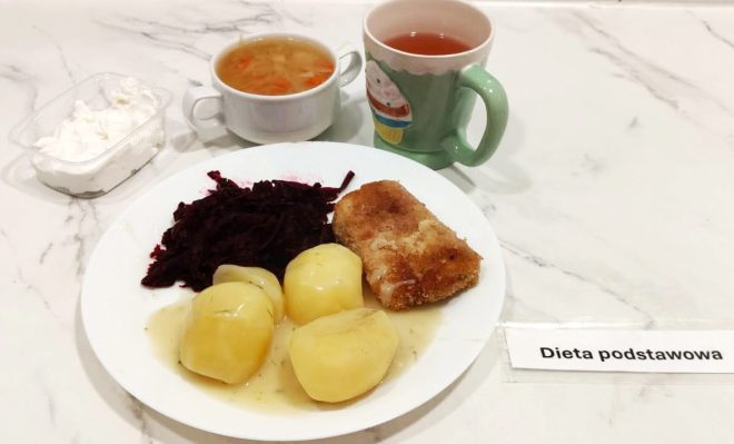 Dieta podstawowa – obiad Talerz z trzema ziemniakami w sosie, buraczkami i panierowaną rybą. W tle miseczka z zupą jarzynową, kubek czerwonego napoju oraz pojemniczek z mizerią
