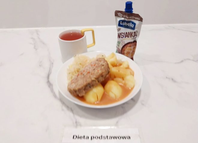 Na talerzu znajduje się kotlet mielony z sosem, ziemniaki oraz surówka z białej kapusty. Do posiłku podano herbatę w białej filiżance oraz deser – owsiankę w tubce marki Lubella. Zestaw opisany jako „Dieta podstawowa”