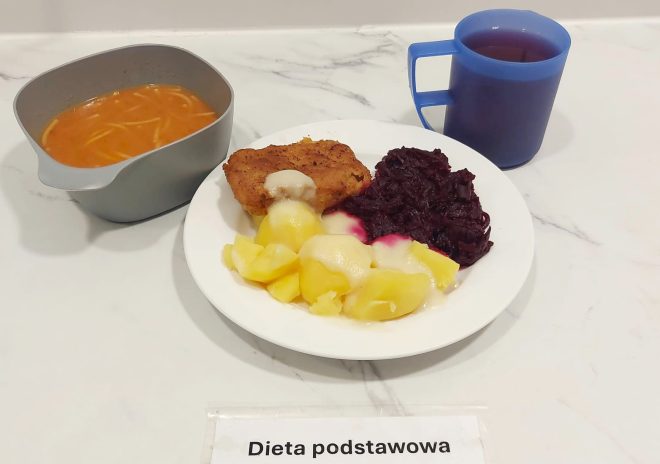 Na talerzu podano gotowane ziemniaki z sosem, panierowany kotlet, buraczki na ciepło oraz porcję białego sosu. Do posiłku dołączono zupę pomidorową z makaronem w szarej miseczce oraz herbatę w niebieskim kubku