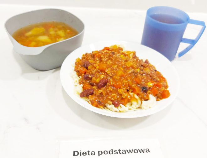 Na talerzu z makaronem podano sos mięsno-warzywny typu bolognese z dodatkiem czerwonej fasoli i kukurydzy. Obok znajduje się miska z klarowną zupą jarzynową zawierającą ziemniaki i marchewkę. Do posiłku dołączono napój w niebieskim kubku. Całość opisana jako „Dieta podstawowa”
