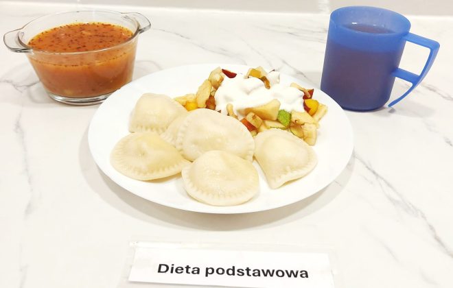 Talerz z kilkoma pierogami, obok porcja sałatki owocowej polanej jogurtem. W tle szklana miseczka z zupą pomidorową i niebieski kubek z kompotem. Na dole kartka z napisem „Dieta podstawowa”