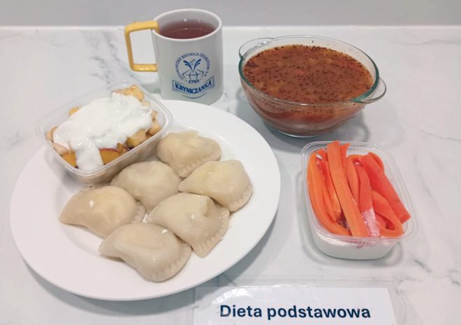 Taca z obiadem w diecie podstawowej: talerz z pierogami ruskimi i sałatką owocową z jogurtem, miska z zupą pomidorową z ziołami, kubek z kompotem oraz porcja marchewek pokrojonych w słupki w plastikowym pojemniku