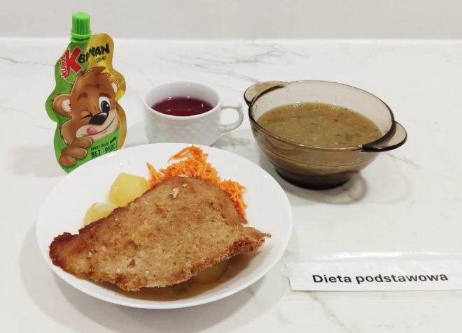 Taca z obiadem w diecie podstawowej: talerz z panierowanym kotletem schabowym, gotowanymi ziemniakami i surówką z tartej marchewki, obok kubek z czerwonym napojem, miska z zupą jarzynową i mus owocowy w saszetce z wizerunkiem misia