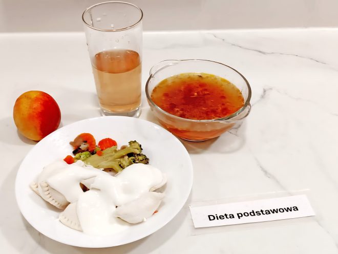 Na białym talerzu podano pierogi polane białym sosem, obok porcję gotowanych warzyw: marchewki i brokułów. Do zestawu dołączono zupę w szklanej miseczce oraz szklankę jasnego napoju. Po lewej stronie leży brzoskwinia. Na blacie znajduje się karteczka z napisem „Dieta podstawowa”