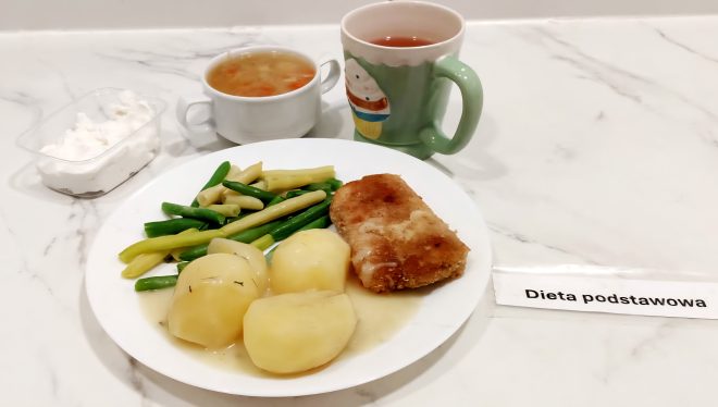 „Dieta podstawowa” Obiad na białym talerzu: gotowane ziemniaki w całości, zielona i żółta fasolka szparagowa, panierowany kotlet schabowy z sosem, obok miseczka zupy jarzynowej, porcja twarogu oraz kubek herbaty w zielonym kubku z motywem babeczki
