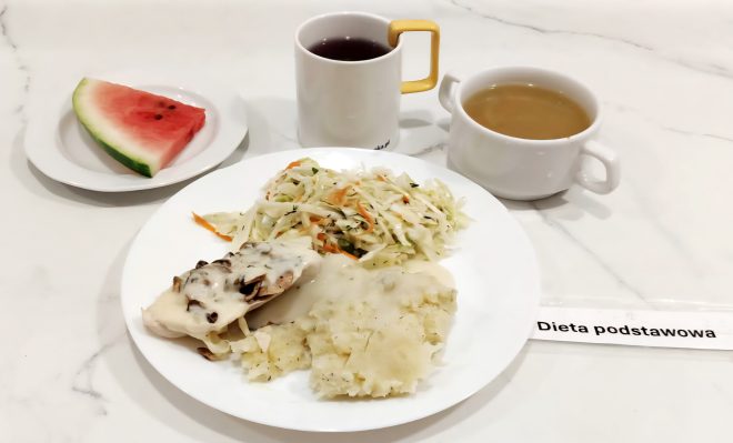 Talerz z puree ziemniaczanym polanym sosem, surówką z białej kapusty i marchewki oraz pieczonym filetem z kurczaka pod sosem pieczarkowym. Obok na osobnym talerzyku trójkątny kawałek arbuza, a w tle kubek z czerwonym napojem i miseczka z bulionem. Posiłek w diecie podstawowej