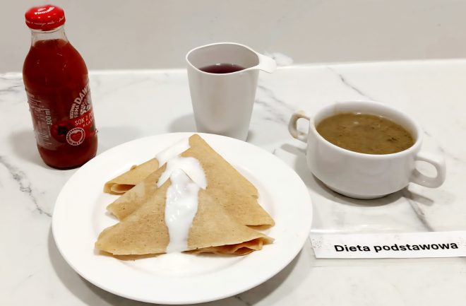 Talerz z trzema naleśnikami polanymi jogurtem naturalnym, obok filiżanka z herbatą, miseczka z zupą pieczarkową i butelka soku z czerwonej porzeczki, etykieta „Dieta podstawowa”