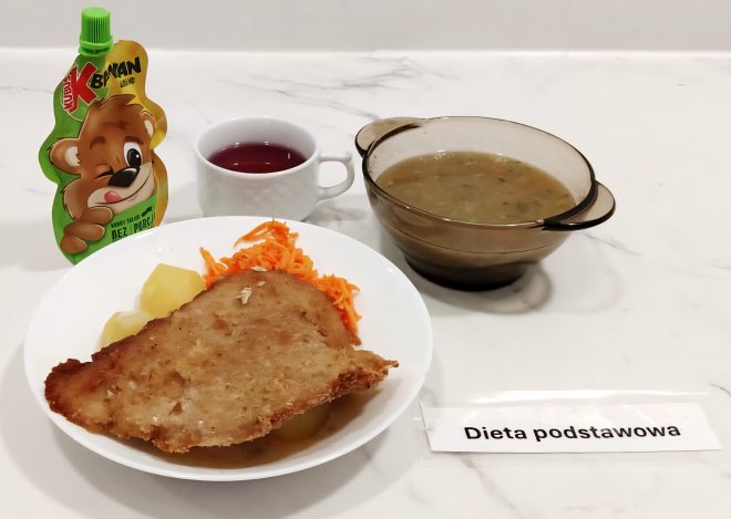 Na białym talerzu podany jest kotlet schabowy panierowany z gotowanymi ziemniakami i surówką z marchewki. Obok znajduje się szklana miseczka z zupą jarzynową, kubek z czerwonym napojem oraz mus owocowy w saszetce z wizerunkiem misia. Na stole leży kartka z napisem „Dieta podstawowa”