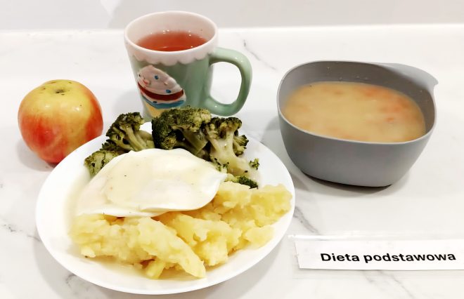 Na stole ustawiono zestaw obiadowy opisany jako „Dieta podstawowa”. Na talerzu znajduje się porcja gotowanych ziemniaków, brokuły oraz kawałek mięsa polanego jasnym sosem. Obok stoi czerwone jabłko oraz kubek z herbatą w zielonym kubku z ilustracją babeczki. Po prawej stronie podano miskę z zupą jarzynową o klarownej konsystencji z kawałkami warzyw
