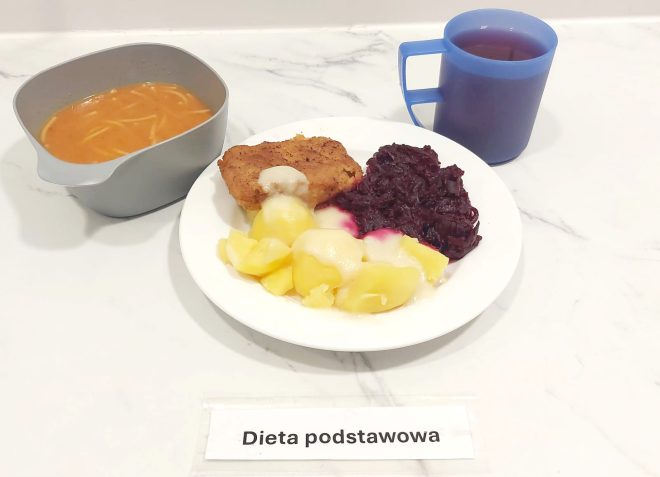 Obiad w diecie podstawowej: na talerzu podano gotowane ziemniaki polane sosem, panierowany kotlet oraz duszoną czerwoną kapustę. Do zestawu serwowana jest zupa pomidorowa z makaronem w szarej miseczce oraz herbata w niebieskim kubku