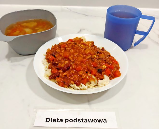Porcja makaronu z sosem mięsno-warzywnym typu bolognese, podana w białym talerzu. Obok kubek z kompotem i miseczka z zupą jarzynową. Podpis: dieta podstawowa