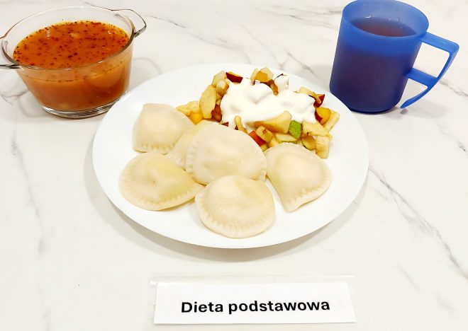 Na białym talerzu ułożono pięć pierogów gotowanych oraz porcję sałatki owocowej z kawałkami jabłka i gruszki, polanej jogurtem naturalnym. Obok stoi miseczka z zupą pomidorową z makaronem oraz napój w niebieskim kubku. Zestaw oznaczono jako „Dieta podstawowa”