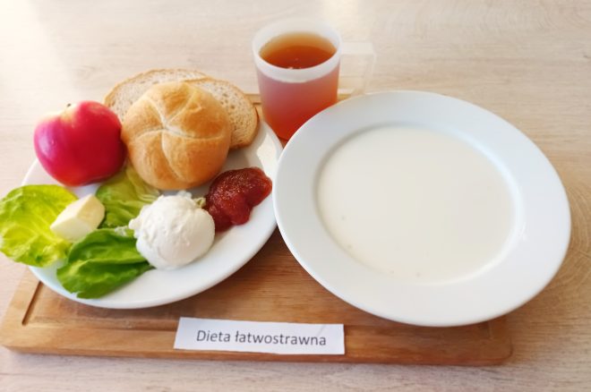 Dieta łatwostrawna: Drewniana taca ze śniadaniem – na talerzu bułka pszenna, kromka chleba pszennego i razowego, jabłko, liście sałaty z masłem, kulka białego twarogu oraz porcja dżemu; obok talerz z jasną zupą mleczną i kubek z ciemnym napojem. Na tacy karteczka z napisem „Dieta łatwostrawna”