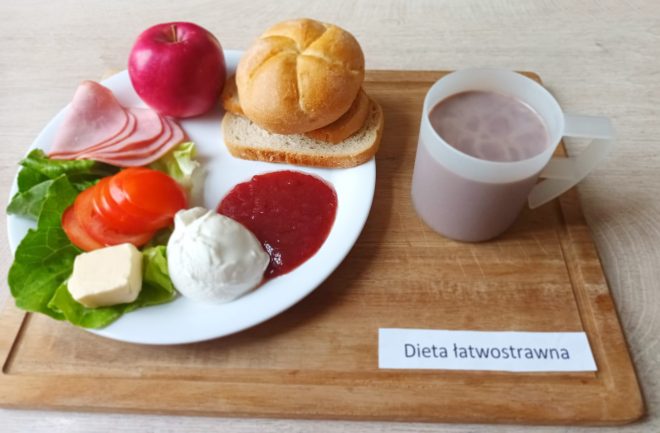 Taca ze śniadaniem w diecie łatwostrawnej: talerz z bułką, kromką chleba pszennego, wędliną, jajkiem na twardo, masłem, plasterkami pomidora, liściem sałaty, porcją dżemu i jabłkiem oraz kubek z kakao