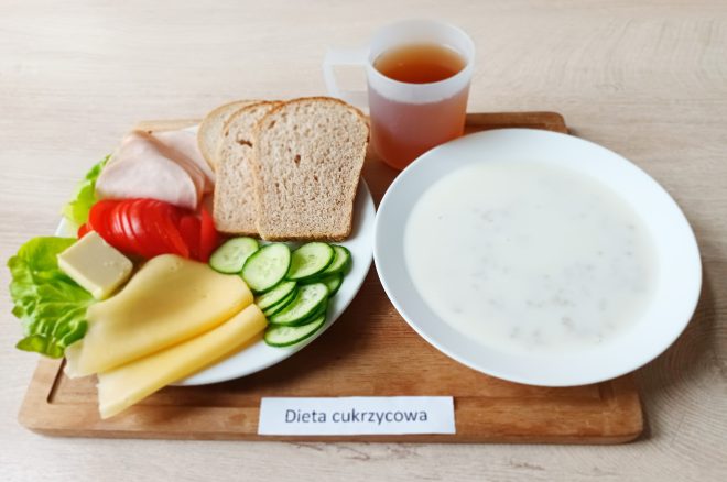 Zdjęcie przedstawia śniadanie w ramach diety cukrzycowej. Na talerzu: dwa plasterki sera żółtego, plasterek sera białego, wędlina drobiowa, plasterki pomidora i ogórka, sałata. Obok dwa kromki chleba razowego. Zestaw uzupełnia kubek z herbatą oraz talerz z mlekiem i płatkami. Całość podana na drewnianej tacy z etykietą „Dieta cukrzycowa”