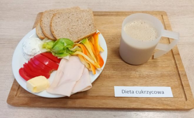 Śniadanie w diecie cukrzycowej na białym talerzu: trzy kromki pieczywa razowego, plasterki pomidora, papryka w trzech kolorach (żółta, czerwona i pomarańczowa), plasterki chudej wędliny z indyka lub kurczaka, liść sałaty, porcja masła oraz kulka serka twarogowego z ziołami. Do posiłku podano napój kawowy z mlekiem lub kawa zbożowa w przezroczystym plastikowym kubku. Całość na drewnianej tacy oznaczonej etykietą: „Dieta cukrzycowa”