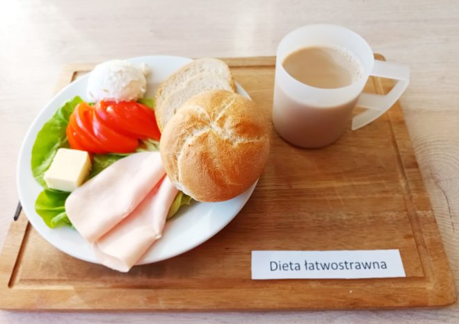 Na białym talerzu ułożonym na drewnianej tacy znajduje się porcja śniadaniowa: bułka kajzerka i kromka jasnego pieczywa, dwa plastry delikatnej wędliny drobiowej, plasterek masła, liść sałaty, kilka plastrów świeżego pomidora oraz kulka białego twarożku. Obok talerza, po prawej stronie, stoi przezroczysty kubek z beżowym napojem, prawdopodobnie kawą z mlekiem. Na tacy umieszczona jest etykieta z napisem „Dieta łatwostrawna”