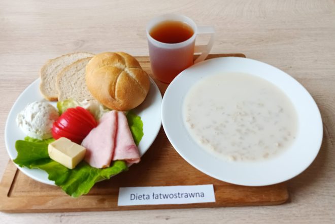Zestaw śniadaniowy w diecie łatwostrawnej: talerz z bułką i kromką chleba pszennego, plasterkiem szynki, plastrem żółtego sera, masłem, porcją twarożku ze szczypiorkiem oraz kawałkiem pomidora na liściu sałaty; obok miska z jasną zupą mleczną z płatkami owsianymi i kubek herbaty