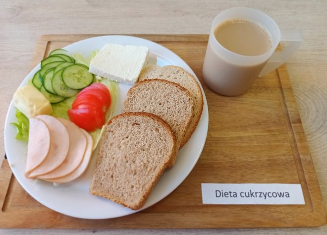 Na białym talerzu znajduje się porcjowany posiłek dietetyczny: trzy kromki chleba pełnoziarnistego, plasterki wędliny drobiowej, ser typu feta, kawałek masła, sałata, plasterki ogórka i pomidora. Obok, w przezroczystym kubku, podano kawę z mlekiem. Posiłek opisano jako „Dieta cukrzycowa”