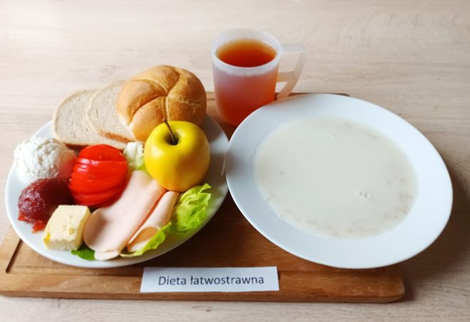 Na drewnianej tacy stoi zestaw śniadaniowy opisany jako „Dieta łatwostrawna”. Po lewej stronie na białym talerzu ułożono bułkę, dwa kawałki jasnego pieczywa, plaster żółtego sera, kilka plastrów wędliny z kurczaka lub indyka, porcję białego twarożku, porcję galaretki owocowej, plasterki pomidora, liść sałaty i żółte jabłko. Obok znajduje się przezroczysty kubek z herbatą. Po prawej stronie stoi talerz z zupą mleczną lub kleikiem na mleku