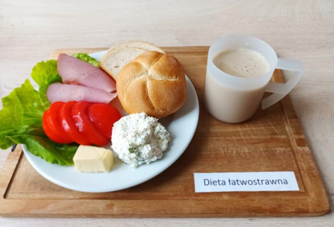 Na drewnianej tacy podano śniadanie w ramach diety łatwostrawnej. Na talerzu leży bułka pszenna i kromka chleba, plasterki wędliny drobiowej, świeże plastry pomidora, liść sałaty, kostka masła oraz porcja twarożku ze szczypiorkiem. Obok ustawiono przezroczysty kubek z jasnym napojem mlecznym