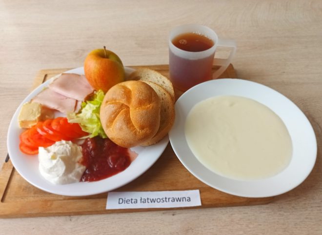 Na zdjęciu przedstawiono posiłek w diecie łatwostrawnej. Na głównym talerzu ułożono świeżą bułkę, kromki białego pieczywa, plasterki szynki drobiowej, pomidora, sałatę, twarożek, ser żółty, porcję dżemu oraz całe jabłko. Obok znajduje się talerz z kremową, białą zupą (np. zupa krem). Do zestawu podano kubek z jasną herbatą. Całość podana na drewnianej tacce z podpisem „Dieta łatwostrawna”