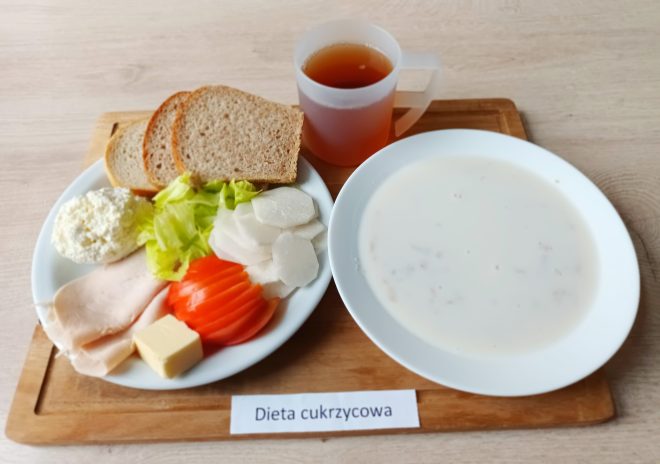 Talerz z kromkami pieczywa razowego, plasterkami pomidora i rzodkwi, sałatą, plasterkami wędliny drobiowej, serem żółtym i porcją twarogu. Obok biały talerz z mlekiem i płatkami oraz kubek z herbatą. Na tacy napis: „Dieta cukrzycowa”