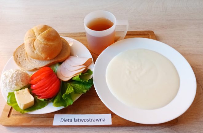Na tacy drewnianej ustawione danie obiadowe opisane jako „Dieta cukrzycowa”. Na talerzu znajduje się porcja makaronu razowego z sosem warzywno-mięsnym w pomarańczowym kolorze oraz surówka z kapusty na liściu sałaty. Obok talerza stoi miska z zupą jarzynową z natką pietruszki, a w przezroczystym kubku podany jest różowy napój owocowy