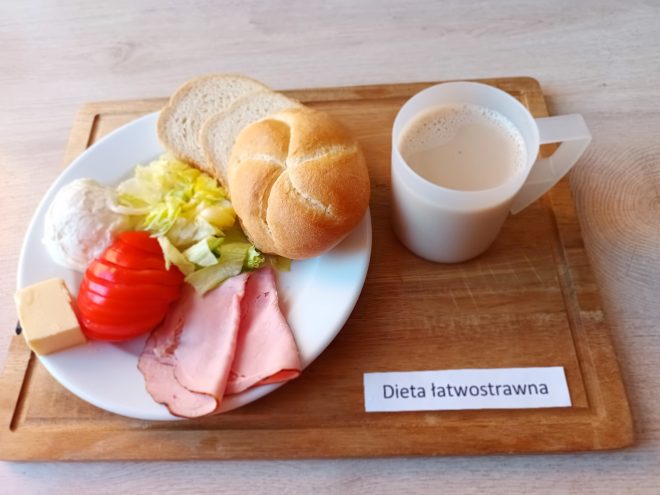 Talerz z pieczywem pszennym – bułką i kromkami chleba, plasterkiem sera żółtego, plastrem pomidora, liściem sałaty, plastrami szynki i kulką twarożku. Do tego biały kubek z kawą z mlekiem lub zbożową. Na tacy napis: „Dieta łatwostrawna”