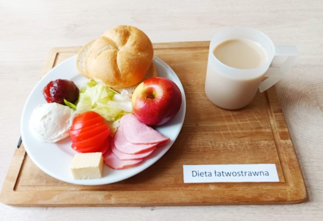 Zestaw śniadaniowy w diecie łatwostrawnej: talerz z bułką, szynką, plasterkami pomidora, sałatą, twarożkiem, kawałkiem żółtego sera, porcją dżemu i jabłkiem, obok kubek kawy z mlekiem