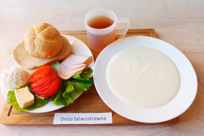 Taca z śniadaniem w diecie łatwostrawnej. Na talerzu bułka i kromka chleba pszennego, plasterki wędliny z indyka, sałata, plasterki pomidora, twarożek, masło. Obok biały talerz z kremową zupą mleczną lub kaszką. W tle kubek z herbatą owocową