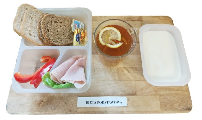 Na drewnianej tacy ułożono śniadanie w plastikowych pojemnikach: w większym trzy kromki pieczywa pszenno‑żytniego, kostkę masła, liść sałaty, dwa plasterki szynki i pasek czerwonej papryki. W mniejszym pojemniku znajduje się gęsty jogurt naturalny. Do picia w przezroczystym kubku podano herbatę z plasterkiem cytryny. Na tacy umieszczono napis „Dieta podstawowa”