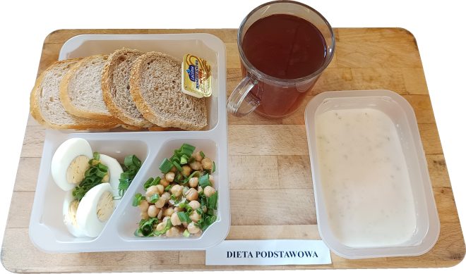 Na tacy podano śniadanie w ramach diety podstawowej. Zestaw obejmuje kromki pieczywa mieszanego z porcją margaryny, gotowane jajko pokrojone w plastry i posypane szczypiorkiem, sałatkę z ciecierzycy z dodatkiem zielonej cebulki, kubek z herbatą oraz pojemnik z jogurtem naturalnym. Wszystkie elementy posiłku są starannie rozmieszczone w przegródkach pojemnika i osobnych naczyniach, a przed nim widnieje karteczka z nazwą diety