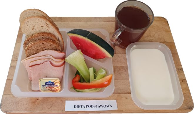 Zestaw śniadaniowy w diecie podstawowej podany na desce: kilka kromek chleba pszennego i razowego, plastry wędliny, porcjowane masło, kawałek arbuza oraz świeże warzywa (ogórek, papryka, seler naciowy). Obok pojemnik z jogurtem naturalnym i szklanka herbaty. Na desce widoczny napis Dieta podstawowa