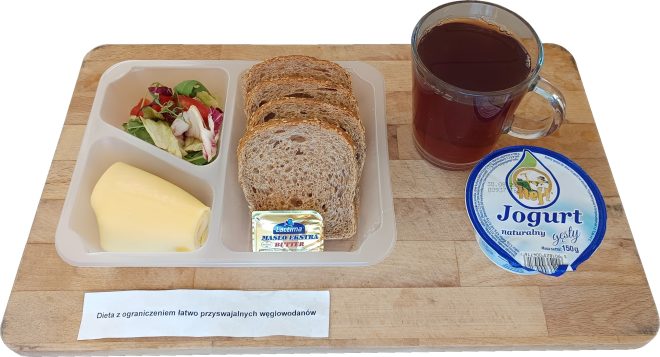 Tacka z pieczywem pełnoziarnistym, masłem i plastrem sera żółtego, porcją mieszanej sałaty. Obok kubek z herbatą oraz jogurt naturalny gęsty w kubeczku. Podpis: „Dieta z ograniczeniem łatwo przyswajalnych węglowodanów”