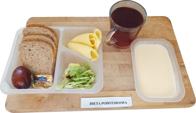 Na tacy znajduje się zestaw: trzy kromki chleba pełnoziarnistego, dwa plastry żółtego sera, masło, śliwka oraz porcje warzyw (ogórek i sałata). Obok kubek z herbatą i miseczka z kaszką mleczną. Podpis na tacy: „Dieta podstawowa”