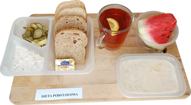 Na tacy drewnianej ułożony zestaw śniadaniowy. W plastikowym pojemniku znajdują się plasterki ogórków kiszonych i sałata oraz pasta twarogowa ze szczypiorkiem. Obok pojemniczek z masłem, trzy kromki chleba wieloziarnistego, szklanka herbaty z cytryną, kawałek arbuza w plastikowym kubeczku i miseczka z jogurtem naturalnym lub owsianką na mleku. Na kartce pod tacą widnieje napis „Dieta podstawowa”
