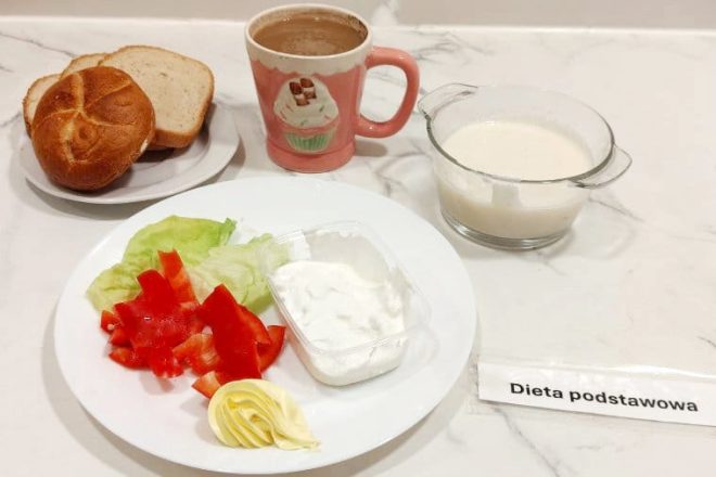 Dieta podstawowa – śniadanie Talerz z liściem sałaty, kawałkami czerwonej papryki, masłem w kształcie róży oraz pojemniczkiem z białym twarożkiem. Obok pieczywo – bułka pszenna i kromki chleba razowego, kubek kawy z mlekiem oraz miseczka z jasną zupą mleczną