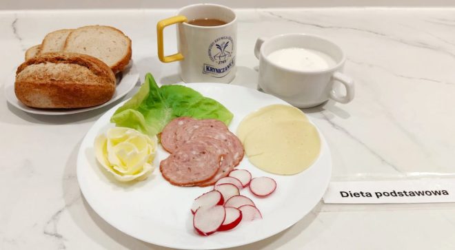 Dieta podstawowa – śniadanie Talerz z liściem sałaty, plasterkami kiełbasy, plastrami sera żółtego, masłem w kształcie róży oraz plasterkami rzodkiewki. Obok pieczywo – bułka pszenna i kromki chleba razowego, filiżanka kawy z mlekiem oraz miseczka z białym jogurtem lub kefirem