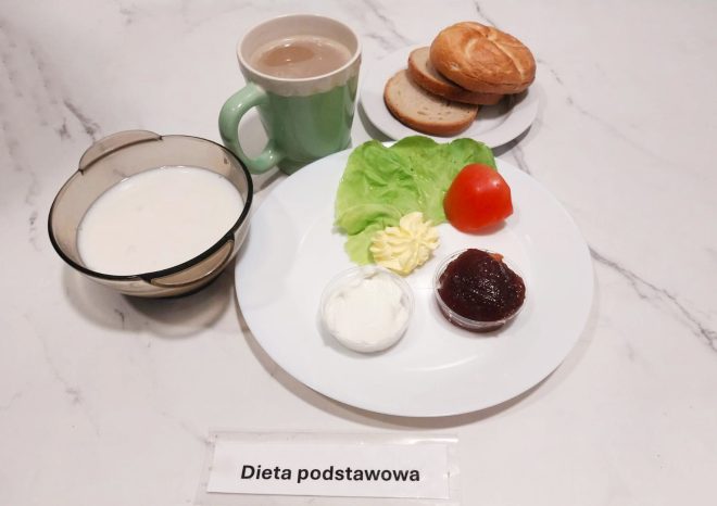 Na talerzu ułożono liść sałaty, plaster pomidora, porcję masła, twarożek oraz konfiturę. Obok stoją kromki chleba i bułka, filiżanka z kawą z mlekiem oraz miseczka z zupą mleczną. Zestaw oznaczony „Dieta podstawowa”