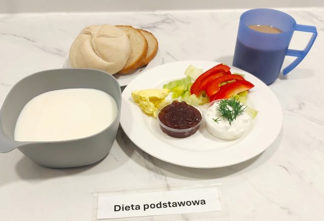 Na talerzu znajduje się sałata, plasterki czerwonej papryki, porcja twarożku ze szczypiorkiem, masło oraz mały pojemniczek z dżemem. W zestawie podano bułkę kajzerkę i kromki chleba, a do picia kawę z mlekiem w niebieskim kubku i mleko w szarej miseczce
